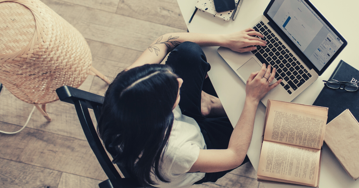 image of a woman typing on a laptop. there are open books and research papers sitting on the desk around her.